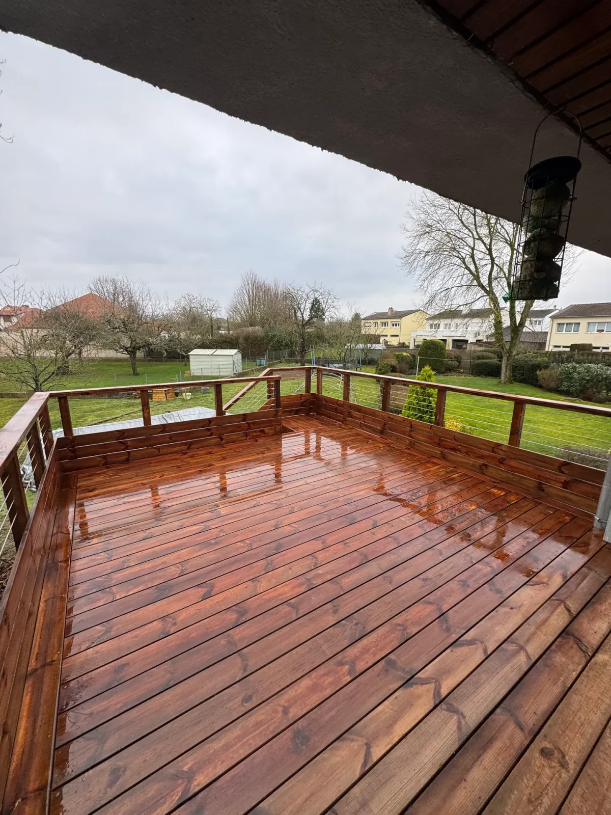 Vue d'une terrasse en bois mouillé avec rampe en bois, dominant un jardin vert avec des arbres nus et des maisons résidentielles visibles au loin sous un ciel gris.