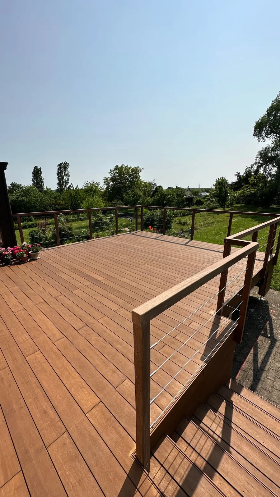 Terrasse en bois clair avec rampe métallique noire, surplombant un jardin avec arbres et pelouse verte par temps ensoleillé.