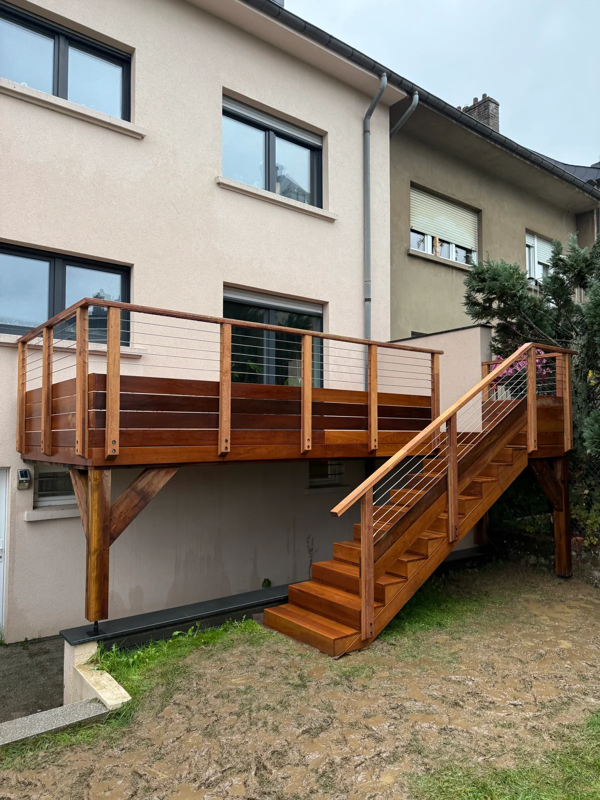 Terrasse en bois massif surélevée avec escalier d'accès et rampe en câbles métalliques, construite contre une maison résidentielle moderne avec fenêtres larges et murs beige et gris.
