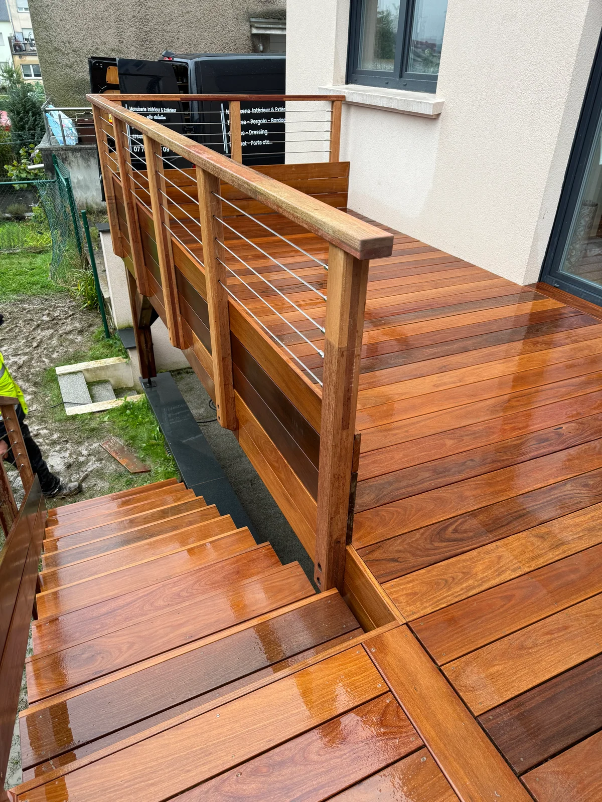 Escalier et terrasse en bois verni de couleur miel, avec rampe en bois et câbles métalliques, menant à une maison contemporaine avec murs blancs et portes-fenêtres noires.